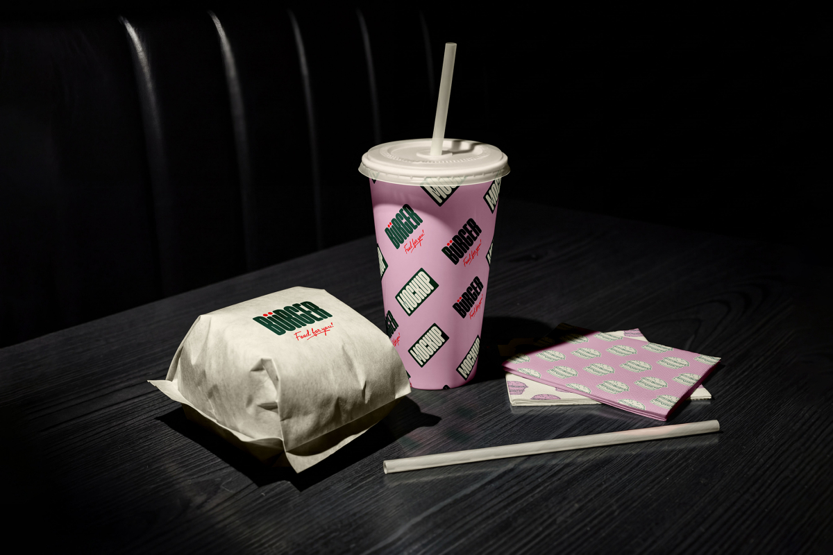 A fast-food meal set arranged on a dark wooden table in dim lighting, showing a burger wrapped in white paper, a pink patterned beverage cup with a white straw, and a stack of pink napkins with a burger print.