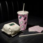 A fast-food meal set arranged on a dark wooden table in dim lighting, showing a burger wrapped in white paper, a pink patterned beverage cup with a white straw, and a stack of pink napkins with a burger print.