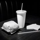 A fast-food meal set arranged on a dark wooden table in dramatic lighting, featuring a burger wrapped in plain white paper, a blank white beverage cup with a straw, a separate straw, and a small stack of plain white napkins.