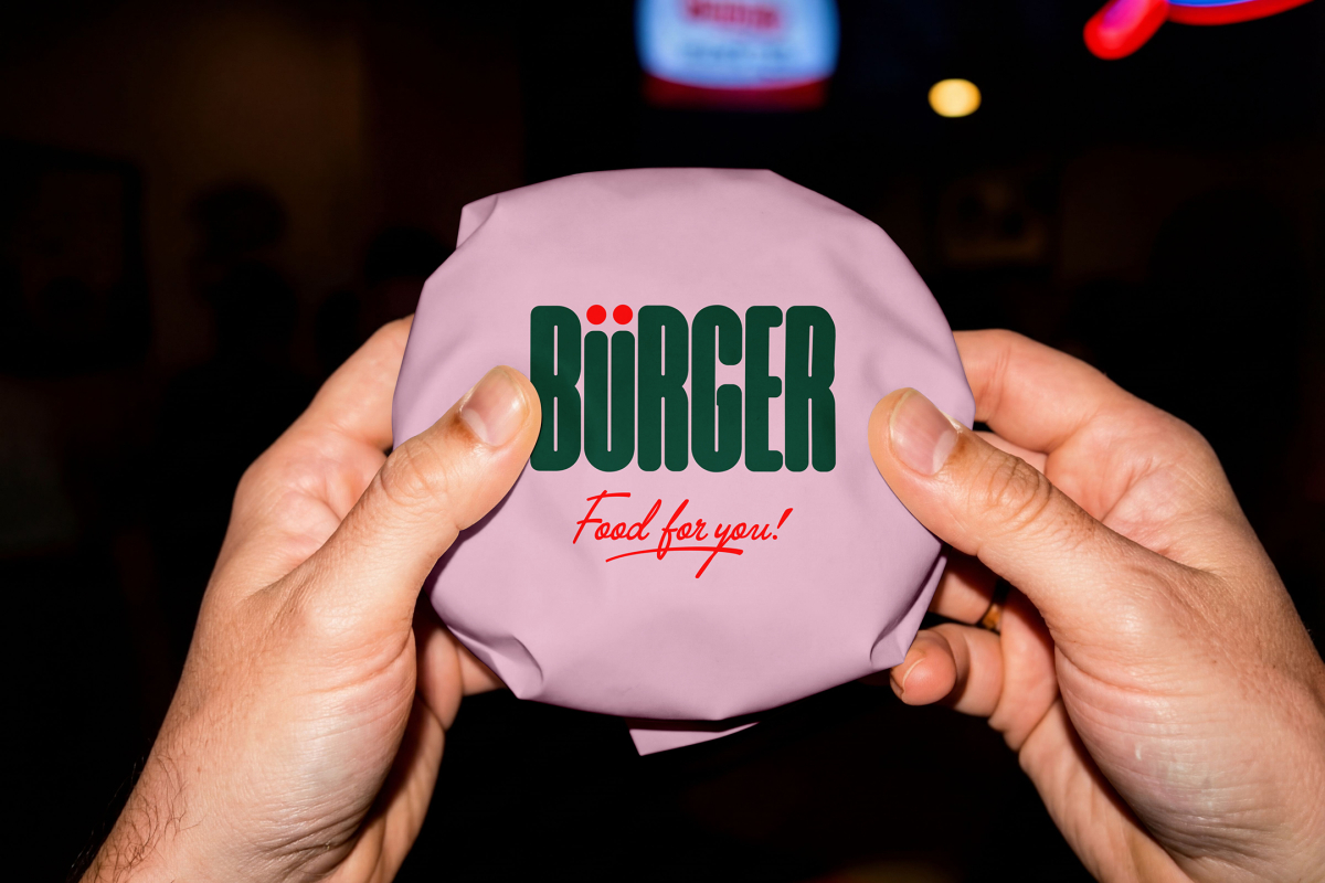 A first-person close-up view of two hands holding a round burger wrapped in light pink paper. The center of the wrapping is printed with a bold green "BÜRGER" logo and a red "Food for you!" tagline against a dimly lit background.