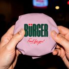 A first-person close-up view of two hands holding a round burger wrapped in light pink paper. The center of the wrapping is printed with a bold green "BÜRGER" logo and a red "Food for you!" tagline against a dimly lit background.