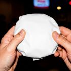 A first-person view of a pair of hands holding a round burger wrapped in plain, unmarked white paper. The background is dimly lit and out of focus.