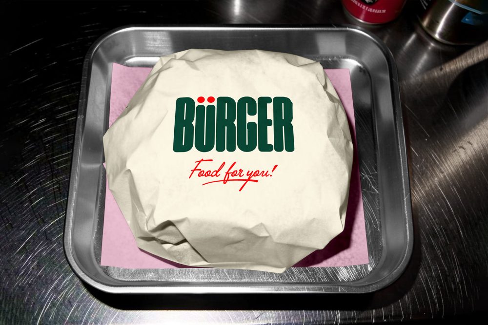 A top-down shot of a burger wrapped in off-white paper, placed on a square sheet of pink tissue paper inside a small metal serving tray. The top of the wrapping displays a large green brand logo and red script text.