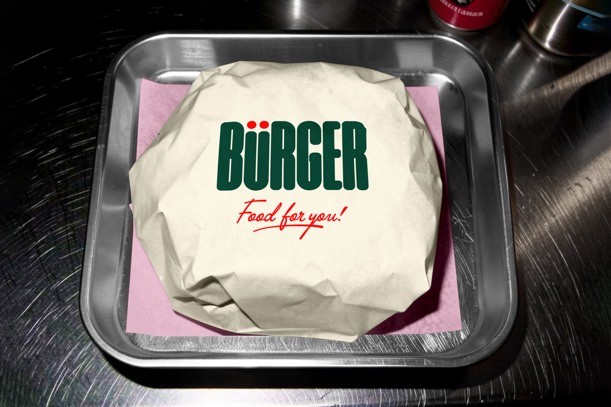 A top-down shot of a burger wrapped in off-white paper, placed on a square sheet of pink tissue paper inside a small metal serving tray. The top of the wrapping displays a large green brand logo and red script text.