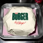 A top-down shot of a burger wrapped in off-white paper, placed on a square sheet of pink tissue paper inside a small metal serving tray. The top of the wrapping displays a large green brand logo and red script text.