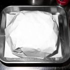 A high-angle, close-up shot of a burger wrapped in blank white paper, sitting on a square piece of white parchment inside a metallic stainless steel serving tray.