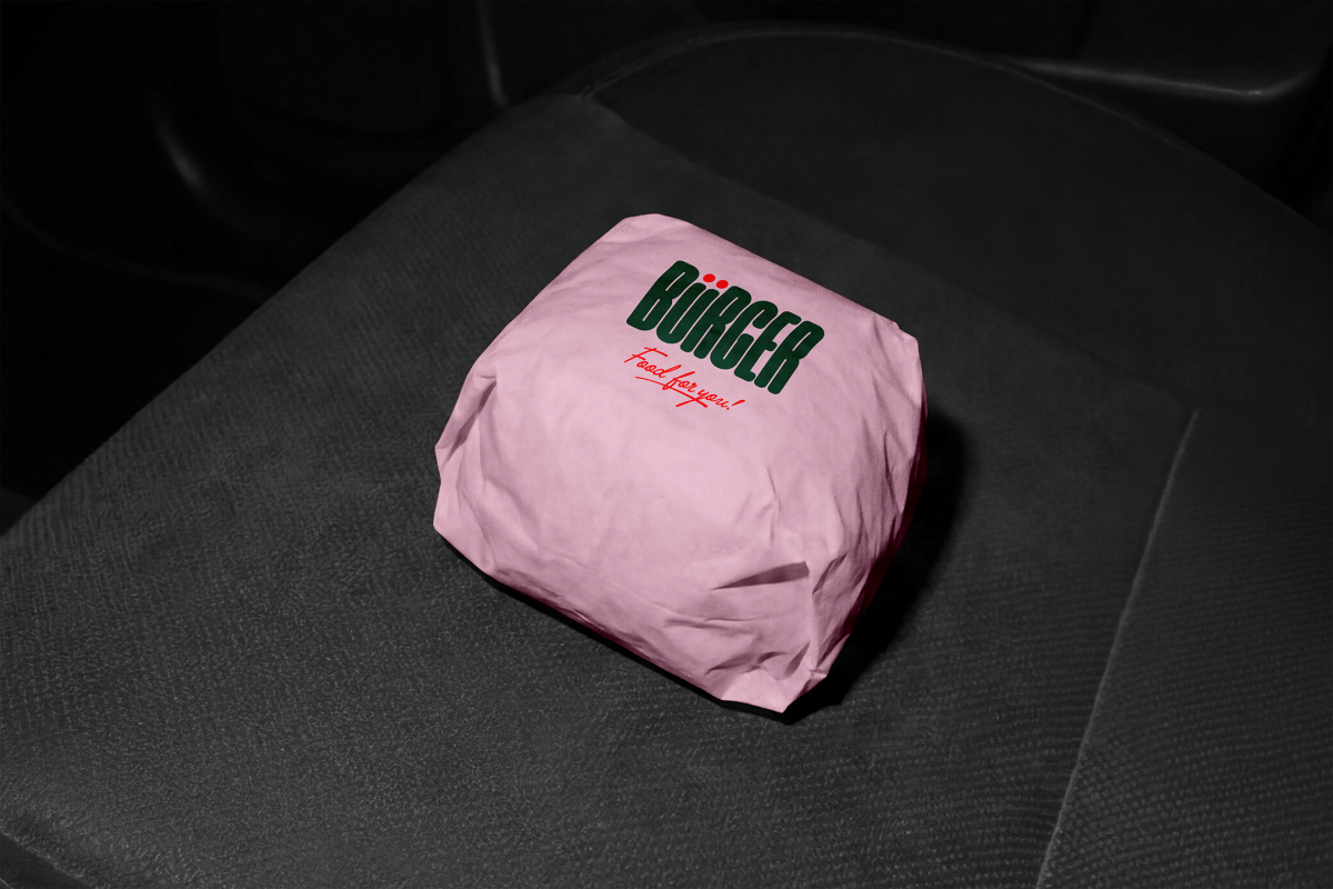 A burger wrapped in pink paper branded with a dark green "BÜRGER" logo and red "Food for you!" text, resting on a dark, textured vehicle seat.