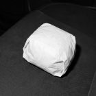 A burger wrapped in plain, unmarked white paper, resting centered on a dark, textured vehicle seat.