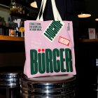 A large light pink canvas tote bag placed on a chrome bar stool inside a restaurant. The bag is printed with a bold green logo, a burger illustration, and red and green "MOCKUP" stickers.