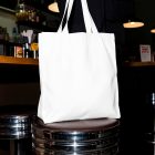 A large, blank white canvas tote bag resting on a chrome-rimmed bar stool with a dark leather top inside a dimly lit restaurant or bar.
