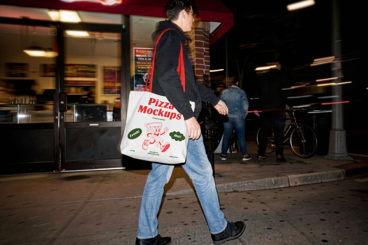 A person walking down a city street at night carrying a white tote bag with red handles. The bag is printed with the "Pizza Mockups" logo, a running pizza slice cartoon, and "Fresh!" and "Hot!" badges.
