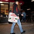 A person walking down a city street at night carrying a white tote bag with red handles. The bag is printed with the "Pizza Mockups" logo, a running pizza slice cartoon, and "Fresh!" and "Hot!" badges.