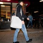 A person walking on a city sidewalk at night in front of a restaurant storefront, carrying a large, blank white canvas tote bag over their shoulder.