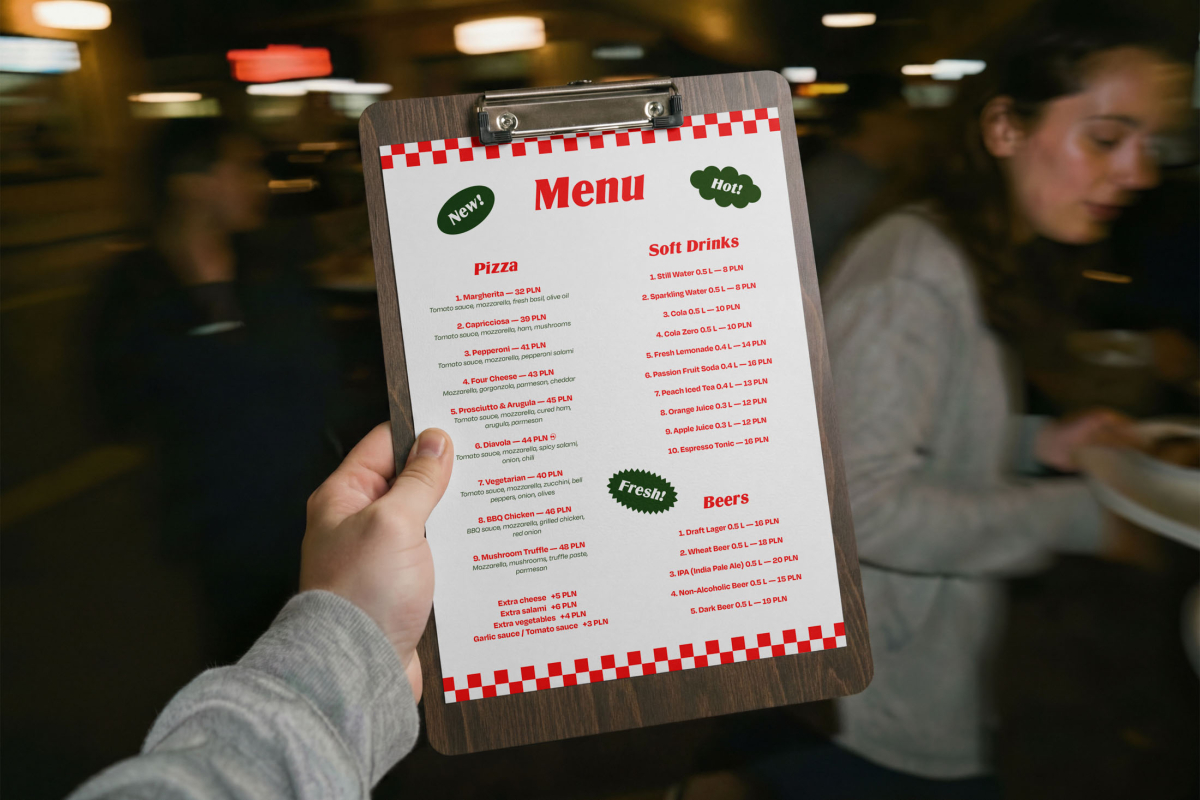 A first-person view of a hand holding a wooden clipboard with a printed restaurant menu. The menu features the "Pizza Mockups" branding, red checkered borders, and lists prices for pizzas, soft drinks, and beers.