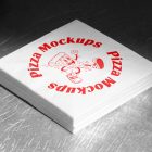 A neat stack of white, square paper napkins sitting on a scratched metal surface. The top napkin is printed with a red circular "Pizza Mockups" logo and a cartoon illustration of a running pizza slice.