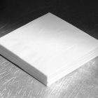 A neat stack of plain white square paper napkins sitting on a brushed and scratched metallic surface under soft light.