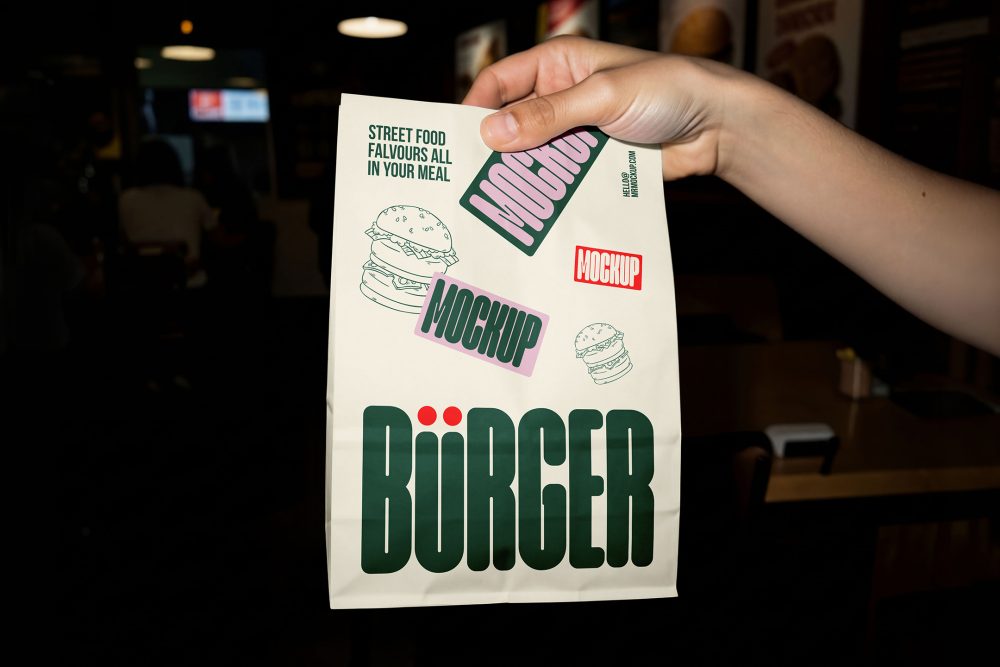 A hand holding up a white paper takeout delivery bag against a dark restaurant background. The bag is decorated with green branding, burger line art, and pink mockup labels.