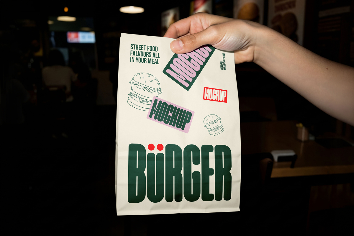 A hand holding up a white paper takeout delivery bag against a dark restaurant background. The bag is decorated with green branding, burger line art, and pink mockup labels.