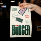 A hand holding up a white paper takeout delivery bag against a dark restaurant background. The bag is decorated with green branding, burger line art, and pink mockup labels.