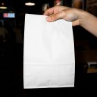 A hand holding up a blank white paper take-out bag against a dark, out-of-focus restaurant background with tables and ambient lighting.