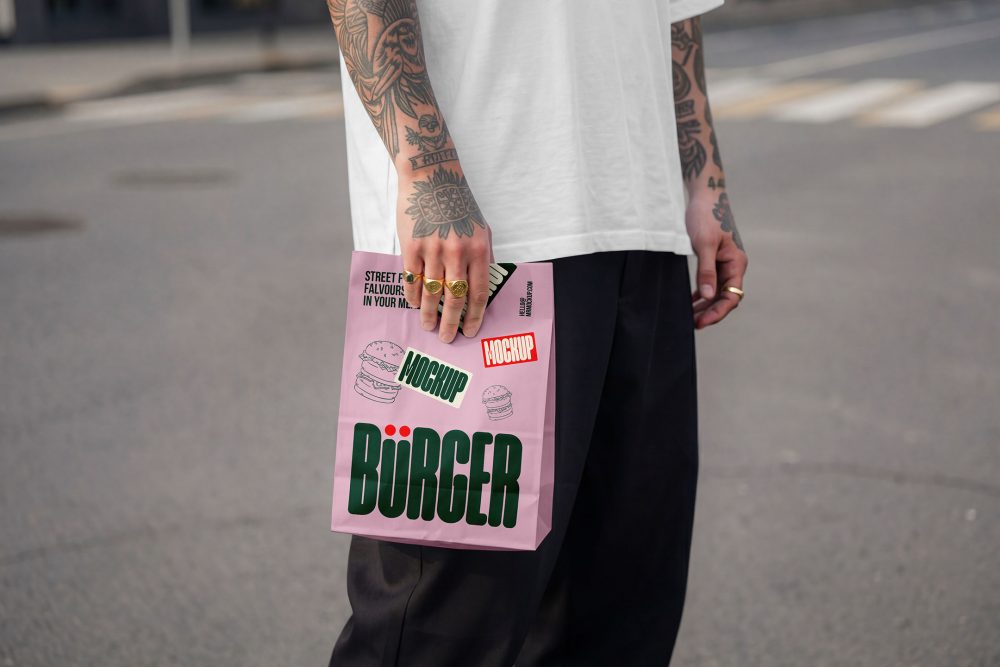 A close-up of a person with heavily tattooed arms wearing a white oversized t-shirt and black trousers, standing on an urban street. They are holding a light pink paper delivery bag decorated with a large green "BÜRGER" logo, burger line art, and mockup badges.