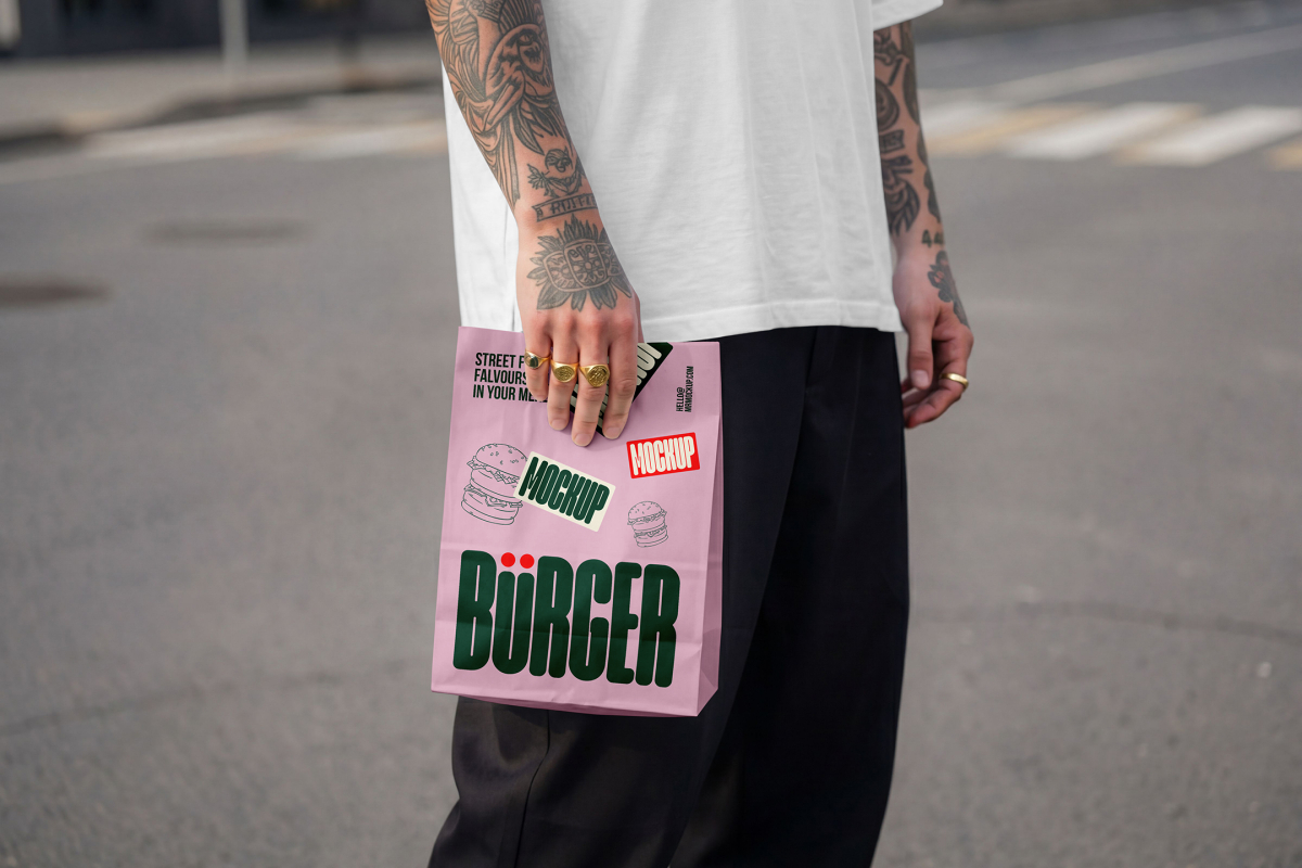 A close-up of a person with heavily tattooed arms wearing a white oversized t-shirt and black trousers, standing on an urban street. They are holding a light pink paper delivery bag decorated with a large green "BÜRGER" logo, burger line art, and mockup badges.