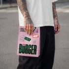 A close-up of a person with heavily tattooed arms wearing a white oversized t-shirt and black trousers, standing on an urban street. They are holding a light pink paper delivery bag decorated with a large green "BÜRGER" logo, burger line art, and mockup badges.