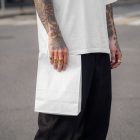A mid-section shot of a person with tattooed arms and gold rings, standing on an outdoor asphalt street. They are holding a plain, blank white paper takeaway bag, wearing a loose white t-shirt and dark trousers.