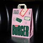 A pink paper shopping bag with white handles placed on a black leather booth inside a restaurant. The bag features a prominent dark green "BÜRGER" logo, burger illustrations, and a bold red "MOCKUP" sticker.