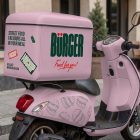 A pastel pink delivery motor scooter parked on a city sidewalk. It features a large matching pink cargo box on the back with "BÜRGER" branding, and a subtle burger line art pattern on the chassis.
