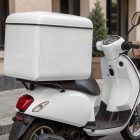 A white delivery scooter parked on a city street pavement. A large, blank white insulated cargo box is mounted on the back rack of the moped.