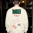 The back view of a person wearing an off-white sweatshirt inside a dimly lit restaurant corridor. The sweatshirt is printed with a dark green "BÜRGER" logo, red script text, and several slanted mockup sticker graphics.