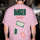 The rear view of a man standing in a commercial restaurant kitchen, wearing an oversized light pink t-shirt. The back of the shirt displays a large green "BÜRGER" logo and three sticker graphics with the text "MOCKUP".