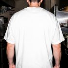 The rear view of a man standing in a commercial kitchen, wearing a blank, oversized white t-shirt. The background shows stainless steel kitchen equipment, shelves with cups, and warm lighting.