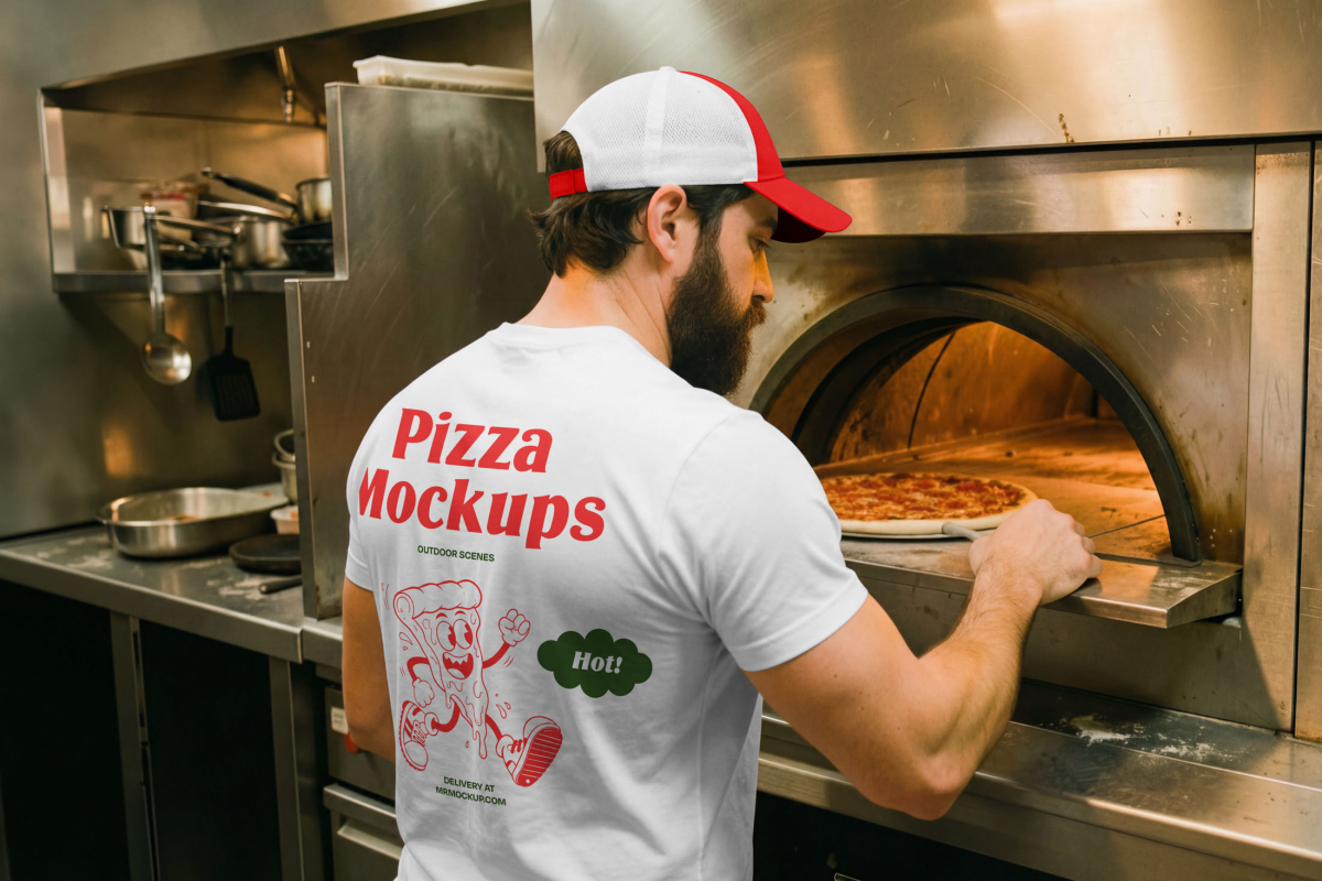 A man wearing a white t-shirt with red "Pizza Mockups" branding on the back and a red and white mesh cap. He is standing in a commercial kitchen, using a pizza peel to check on a baking pizza inside a hot oven.