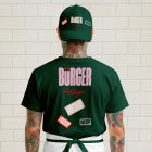 The rear view of a person with neck and arm tattoos standing against a white brick wall, wearing a dark green baseball cap and a matching dark green t-shirt. Both items feature the pink "BÜRGER" logo and various rectangular mockup stickers.