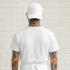 The rear view of a person with colorful tattoos on their neck and arms, standing against a clean white brick wall. They are wearing a blank white baseball cap, a white t-shirt, and a white apron tied at the waist.