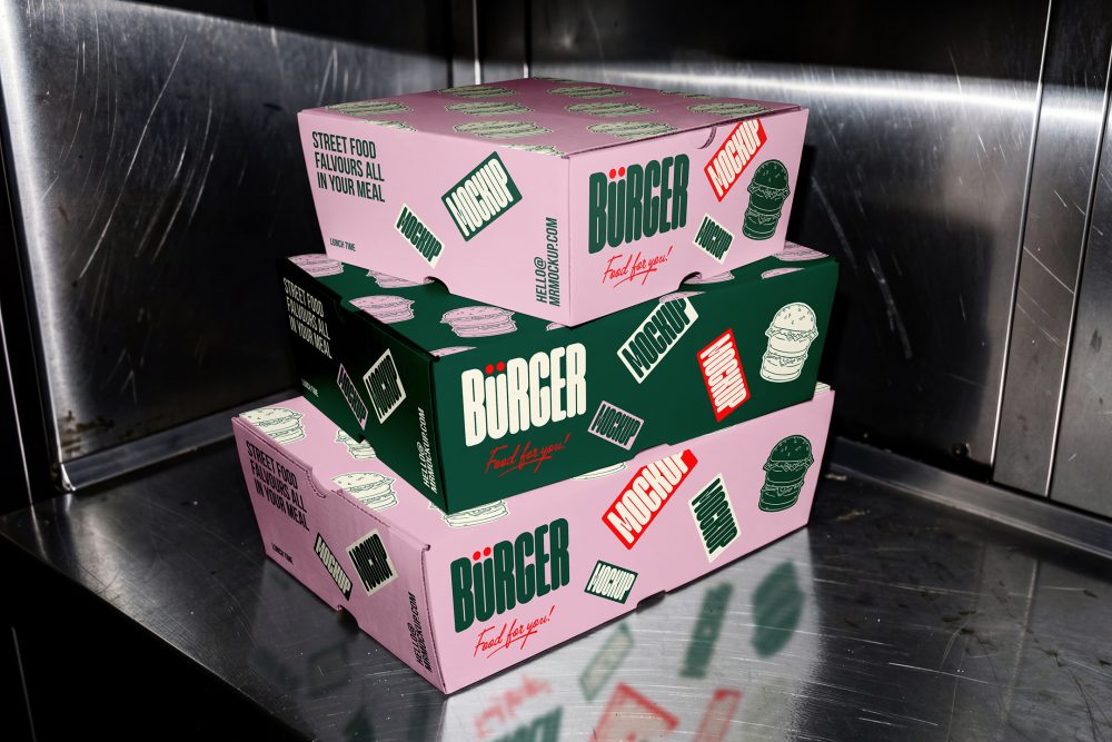 A stack of three burger-style delivery boxes on a stainless steel kitchen counter. The middle box is dark green with white branding, while the top and bottom boxes are light pink with dark green logos and red mockup stickers.