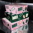 A stack of three burger-style delivery boxes on a stainless steel kitchen counter. The middle box is dark green with white branding, while the top and bottom boxes are light pink with dark green logos and red mockup stickers.