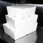 A stack of three plain, unmarked white takeaway cardboard boxes arranged on a scratched stainless steel kitchen counter.