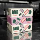 A stack of three rectangular takeout boxes resting on a reflective stainless steel kitchen counter. The middle box is light pink, while the top and bottom boxes are cream-colored. Each box features dark green branding, burger illustrations, and several mockup sticker graphics.