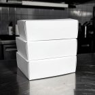 A stack of three blank, unmarked white cardboard takeaway boxes resting on a reflective, brushed stainless steel kitchen counter. The blurred commercial kitchen background features dark tiles and equipment.