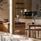 View through a large storefront glass window into a modern, minimal café featuring wooden tables, pour-over coffee apparatus, and warm sunlight.