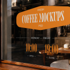 Close-up of a cafe window framed in dark wood with an orange sticker reading "Fresh Coffee Mockups PSD" and opening hours displayed on the glass.