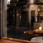 A dark wooden window frame on a café facade reflecting outdoor buildings, showing cozy indoor wooden seating and a small vase on a table.