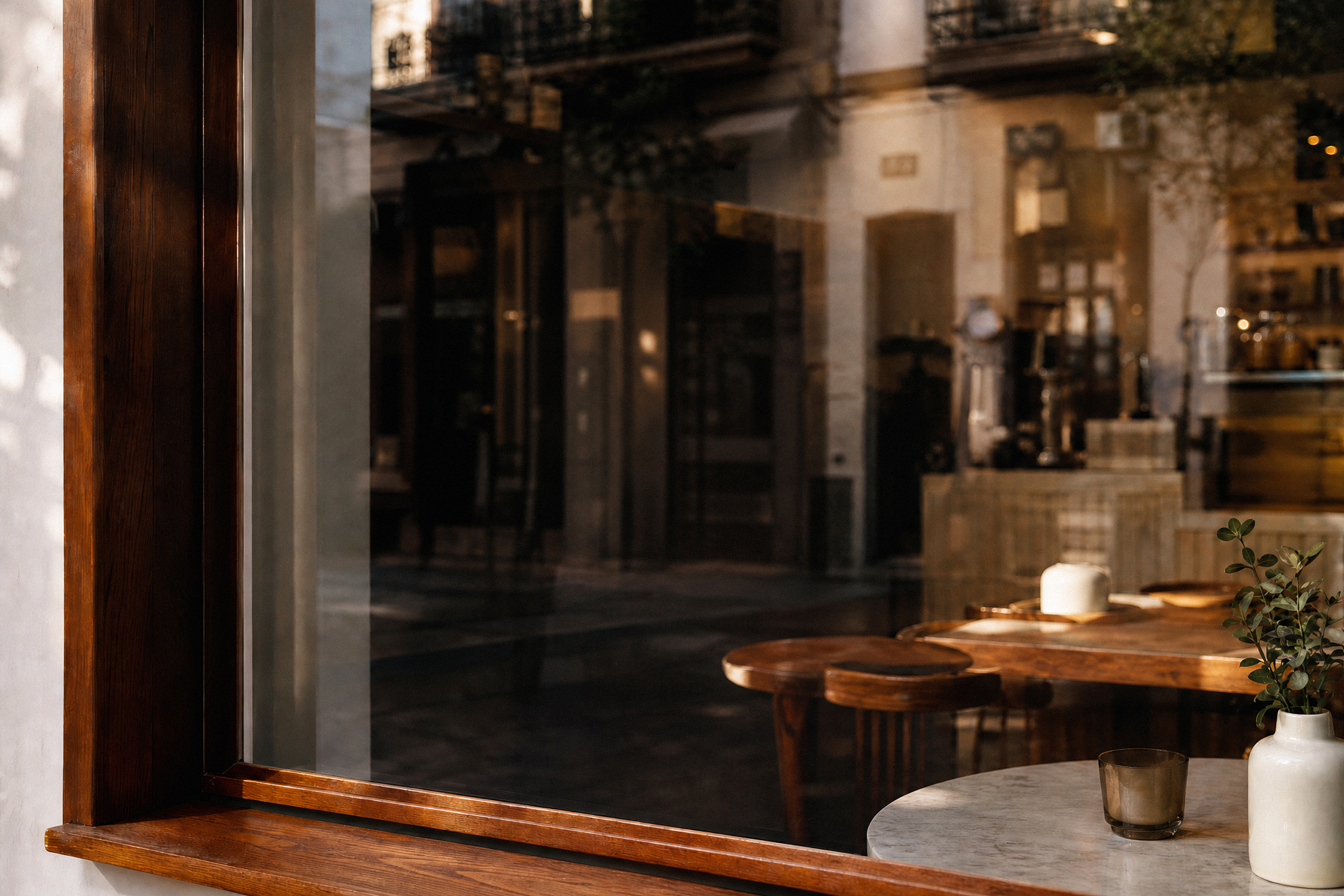 A dark wooden window frame on a café facade reflecting outdoor buildings, showing cozy indoor wooden seating and a small vase on a table.