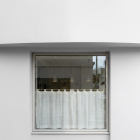 A square window set into a smooth, minimal white wall, covered on the lower half by a white linen cafe curtain, with hanging lights visible inside.