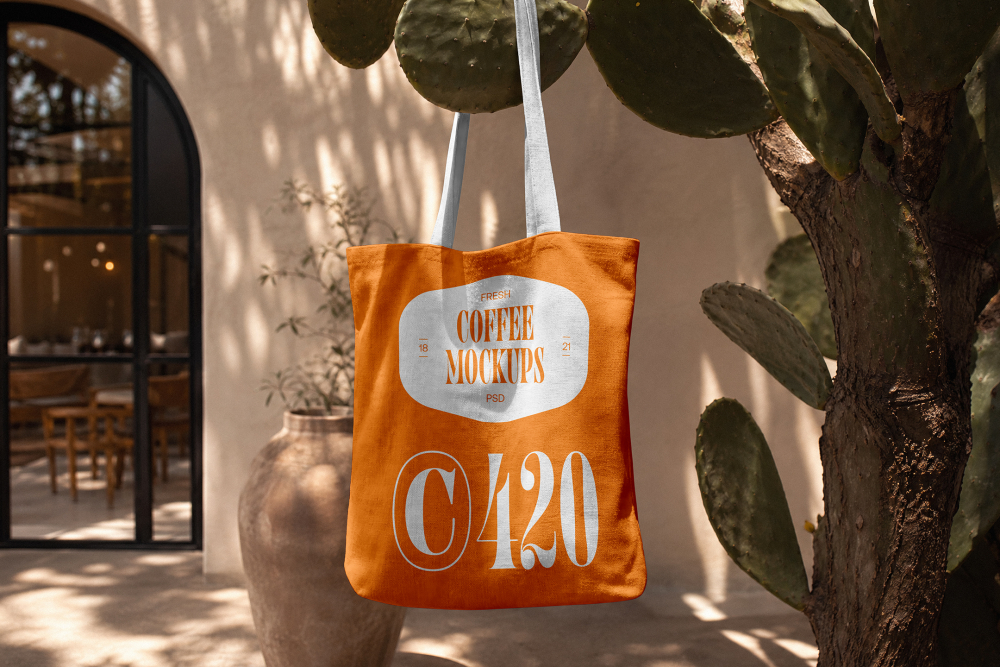 An orange canvas tote bag with white handles hanging from a prickly pear cactus outdoor branch, featuring a white printed coffee logo.