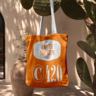 An orange canvas tote bag with white handles hanging from a prickly pear cactus outdoor branch, featuring a white printed coffee logo.