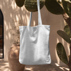 A blank white canvas tote bag hanging from a prickly pear cactus branch in front of a warm-toned Mediterranean building.